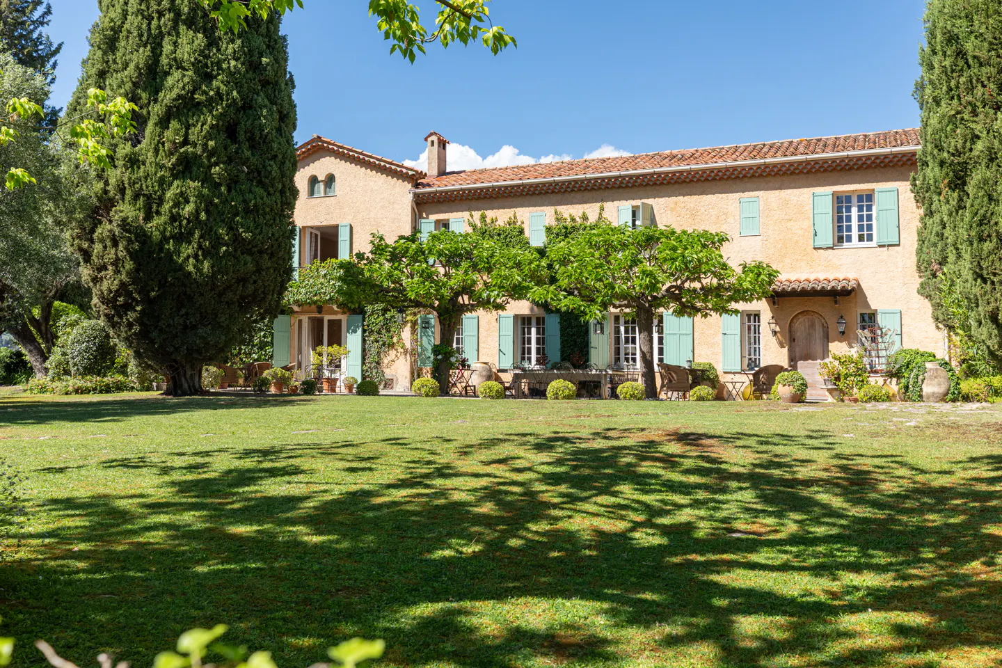 Exterior view of a tan two-story house with green shutters and a red tile roof, surrounded by trees and a green lawn.