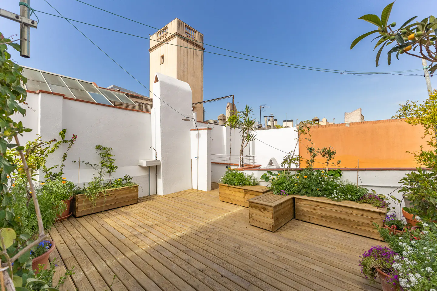A rooftop terrace with a wooden deck, white walls, and planter boxes filled with greenery and flowers under a blue sky.