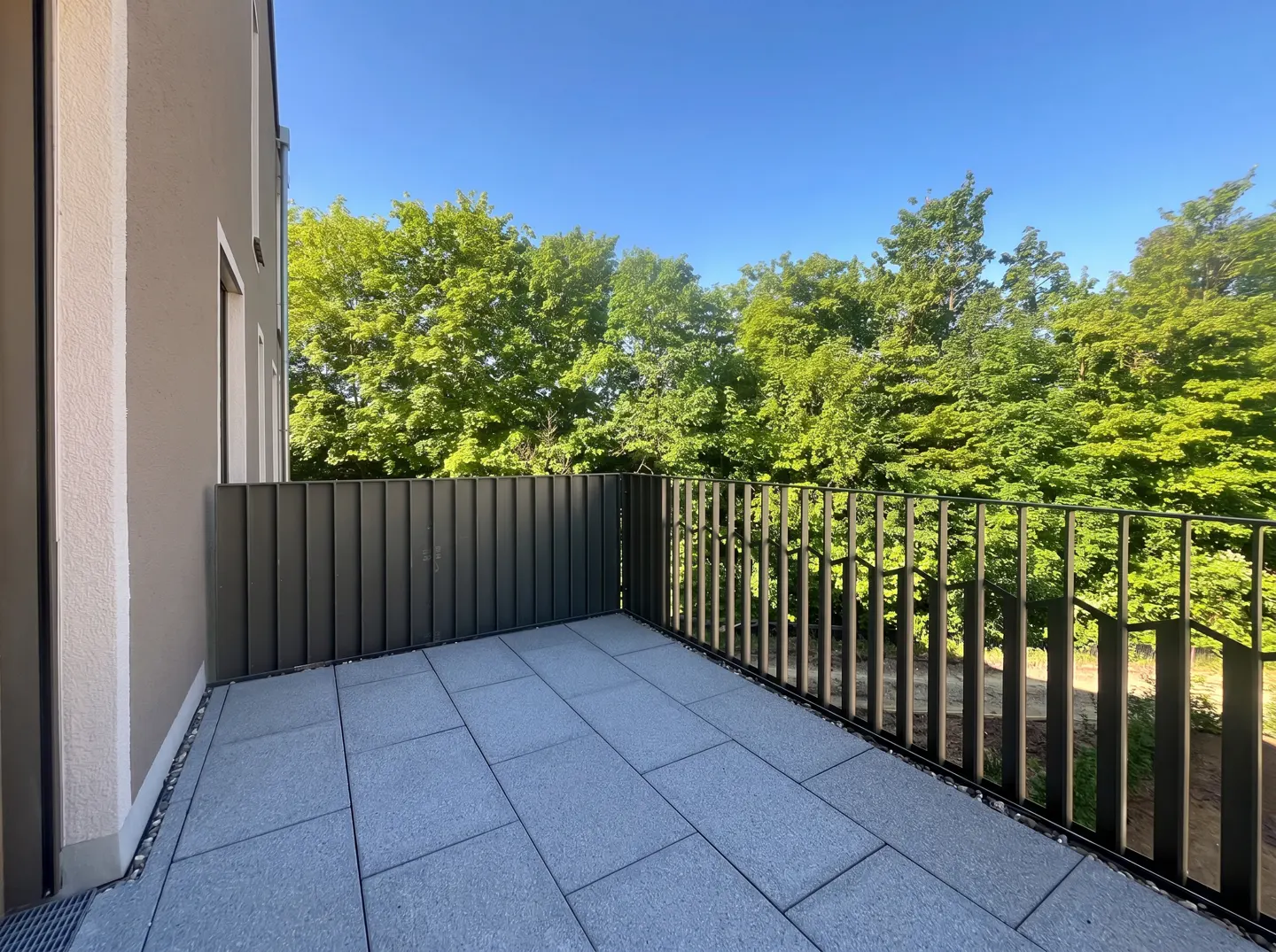 A modern balcony with gray tiles, a dark metal railing, and a view of lush green trees under a clear blue sky.