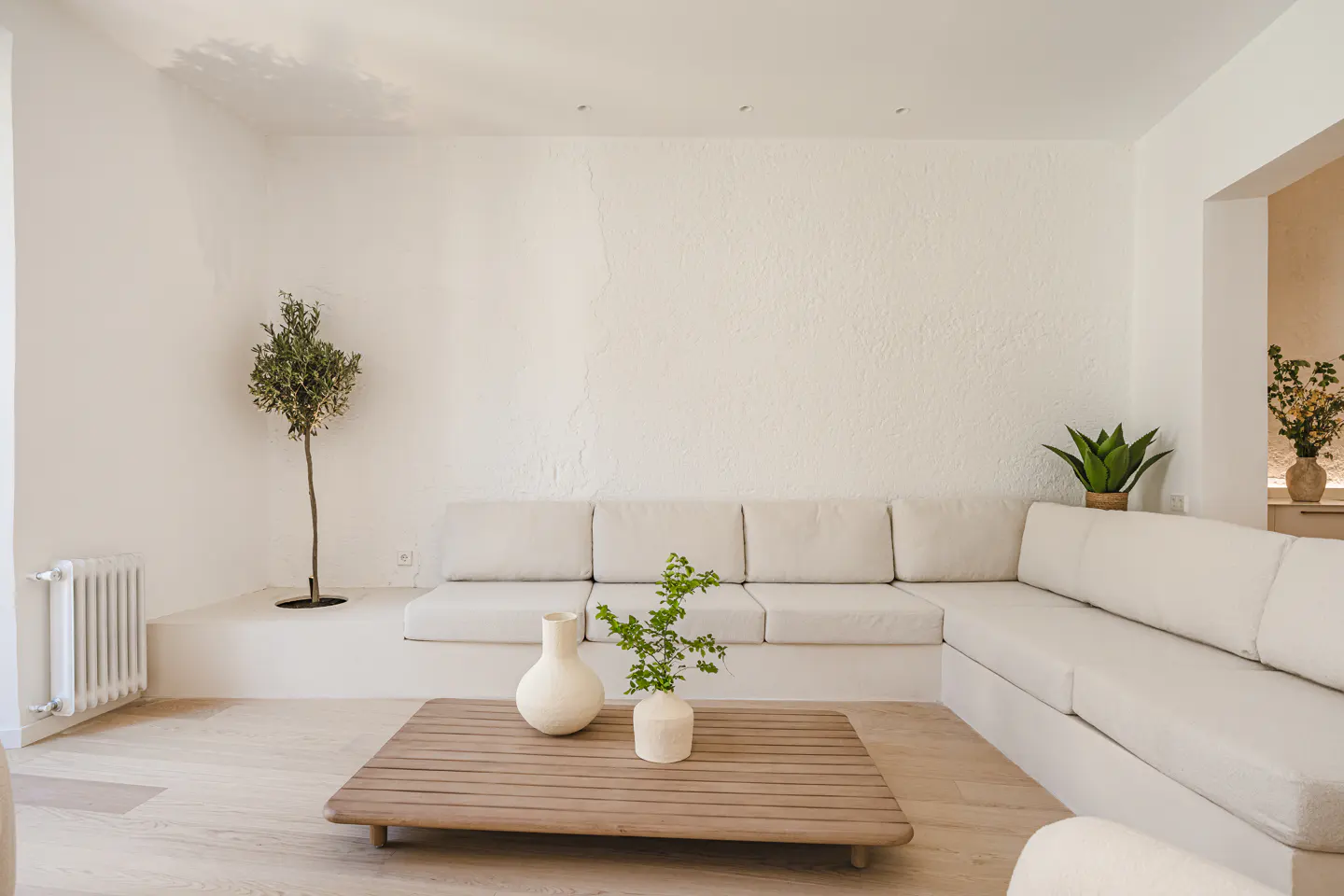 Bright living room with white walls, a beige sectional sofa, and a wooden coffee table with two vases. A small tree and plants add greenery.