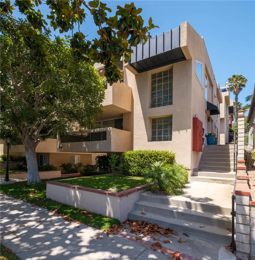 Exterior view of a tan multi-level apartment building with glass block windows, a staircase, and a large tree in front.