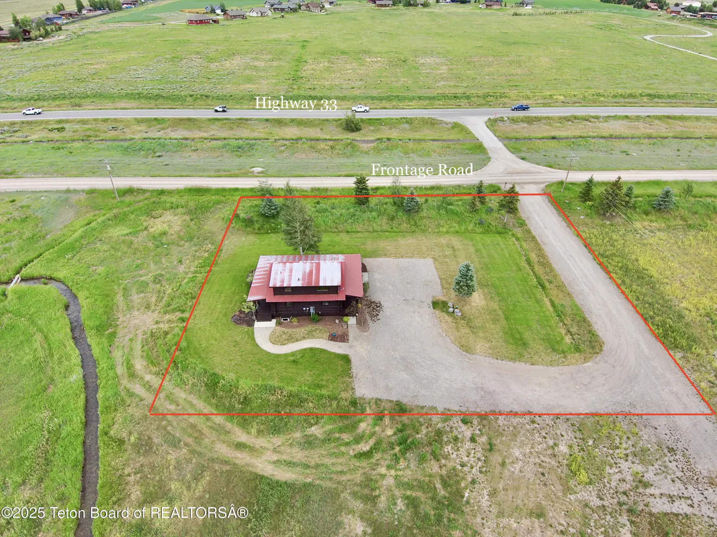 Aerial view of a dark brown house with a red roof, surrounded by green grass and trees, within a red property line. Highway 33 is in the background.