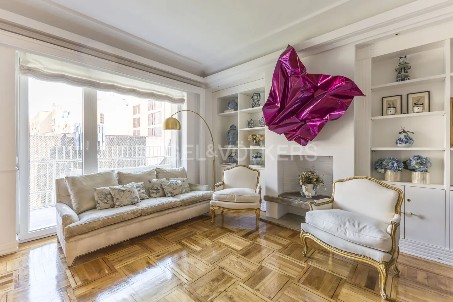 Bright living room with a beige sofa, two white armchairs, and a large pink abstract sculpture above the fireplace. Hardwood floors and built-in shelves.