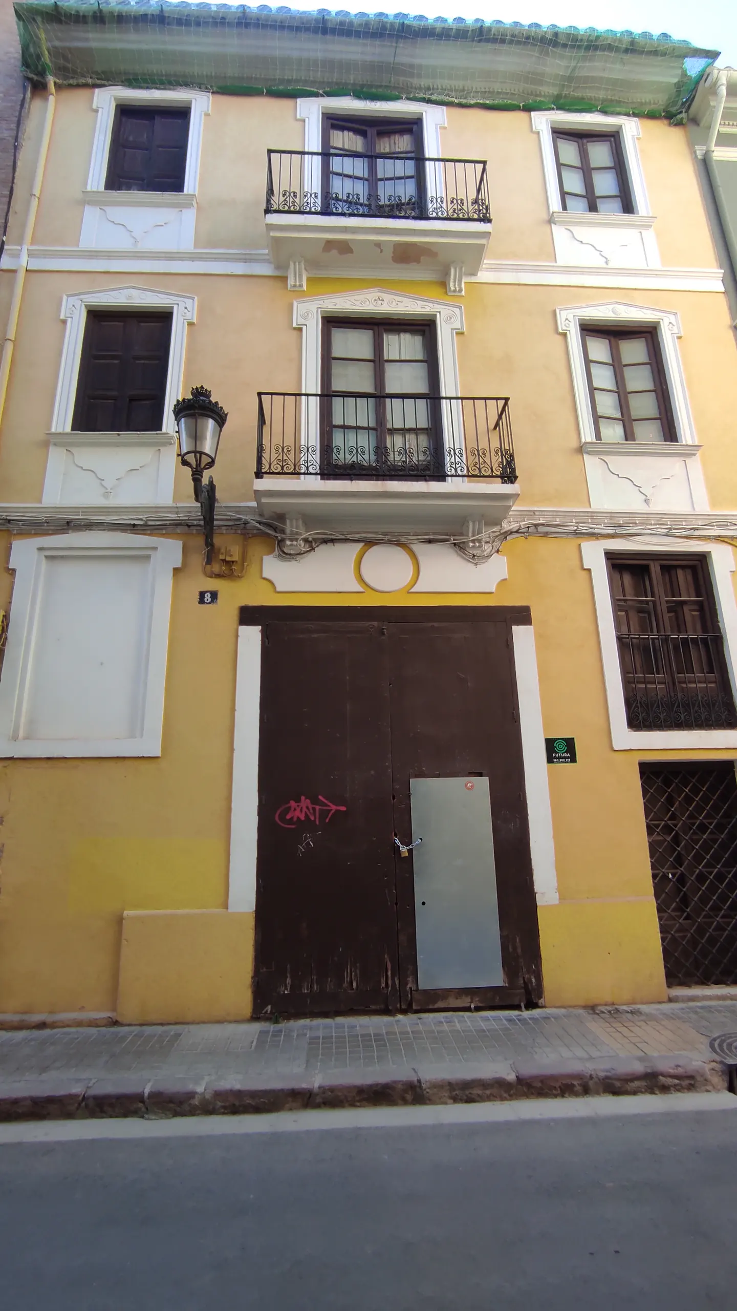 A three-story yellow building with brown doors and window shutters. The building has two small balconies with black railings.