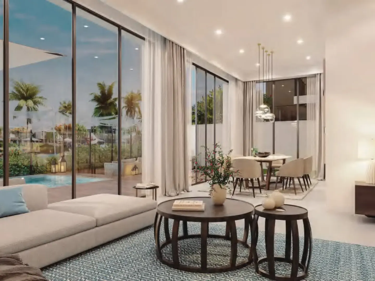 Living room with a beige sofa, round wooden tables, and a dining area with beige chairs. Large windows overlook a pool and palm trees.