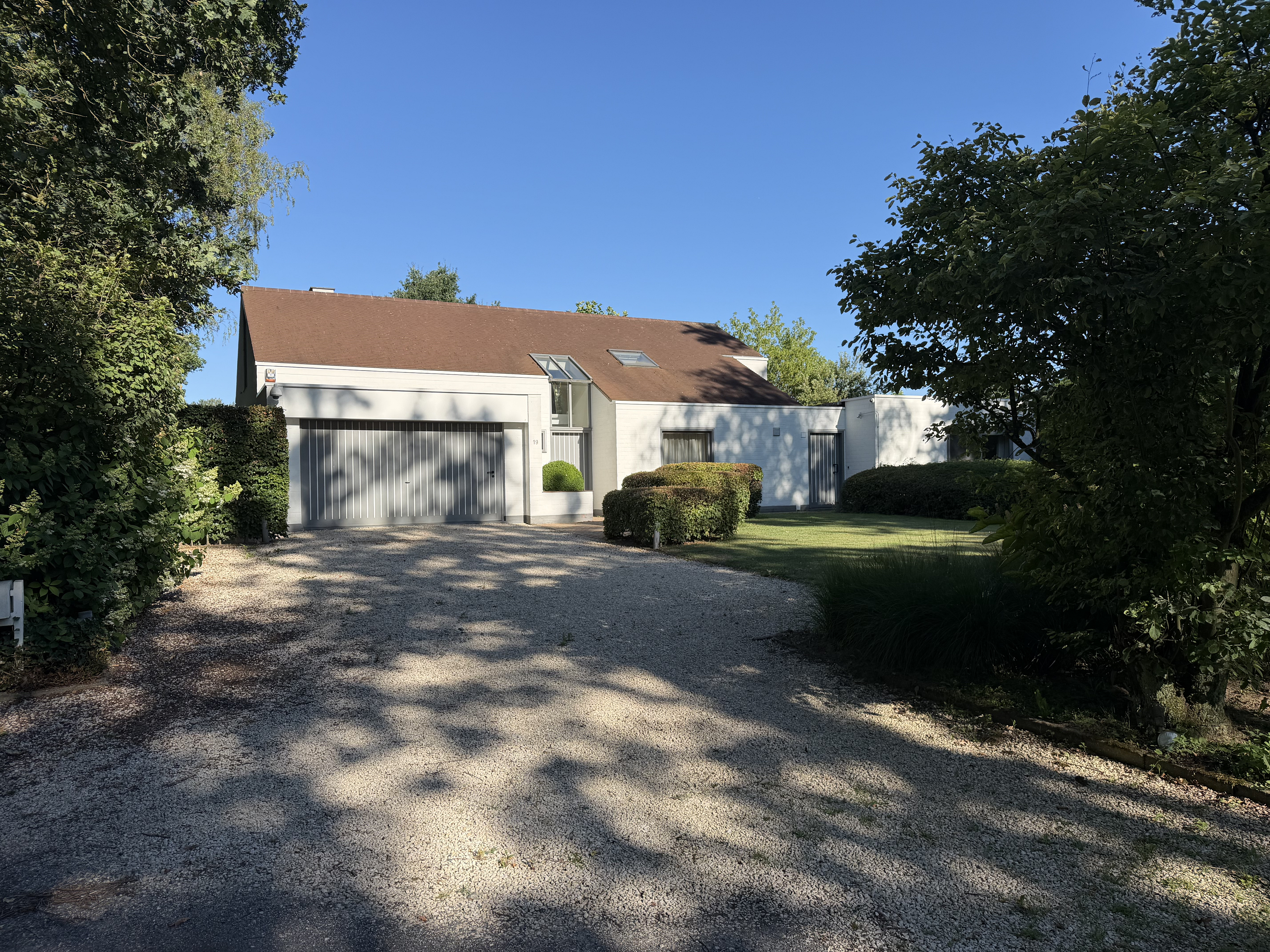 Modern white house with a brown roof, large garage, and gravel driveway, surrounded by lush green trees and landscaping.