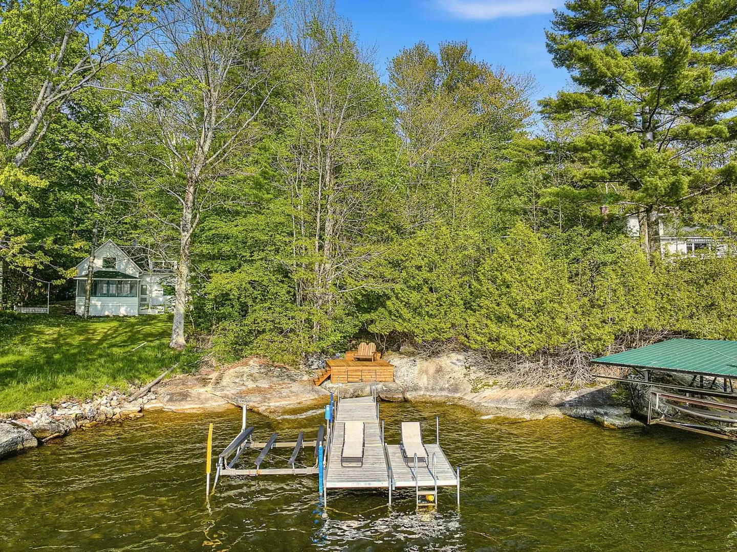 Lakefront property with a dock featuring lounge chairs, surrounded by lush green trees and a small white house in the background.