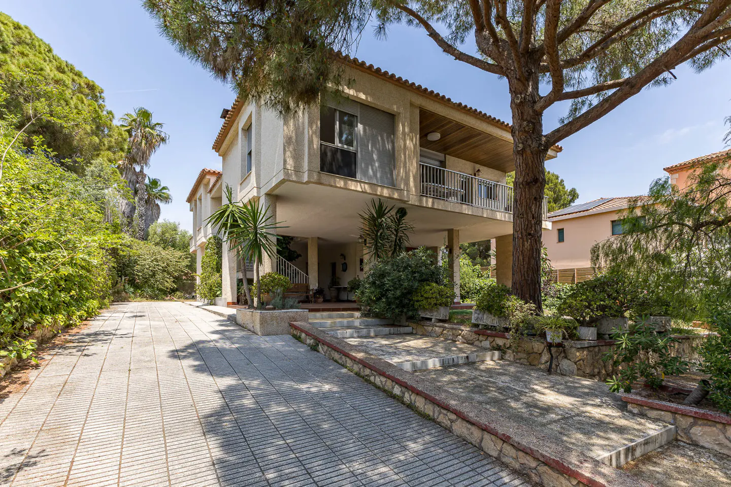 Exterior view of a two-story beige house with a balcony, surrounded by lush greenery and a paved driveway.