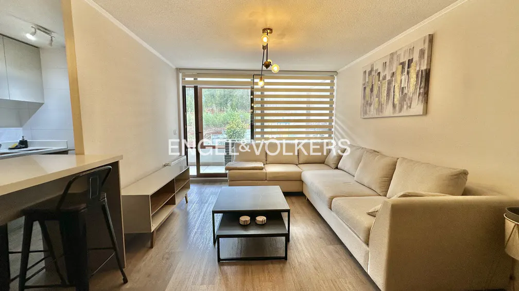 Living room with beige sectional sofa, black coffee table, and light wood floors. A bar with stools separates the kitchen. Balcony with trees visible.