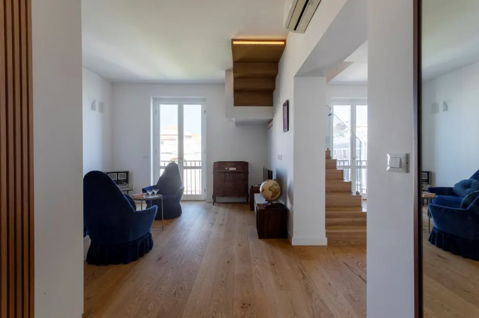 Bright, modern living room with hardwood floors, white walls, and blue velvet chairs. Stairs lead to an upper level. A globe sits on a dark wood cabinet.