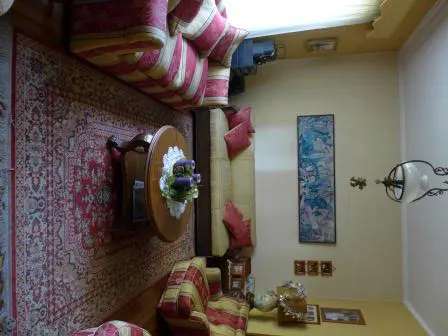A living room with a red patterned rug, striped sofa, and a round wooden table with a floral centerpiece. A painting hangs on the yellow wall.