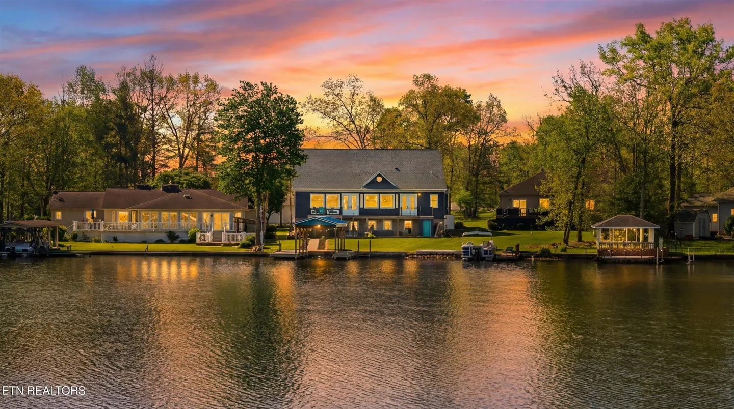 Waterfront homes with docks line a lake under a colorful sunset sky. Trees surround the houses.