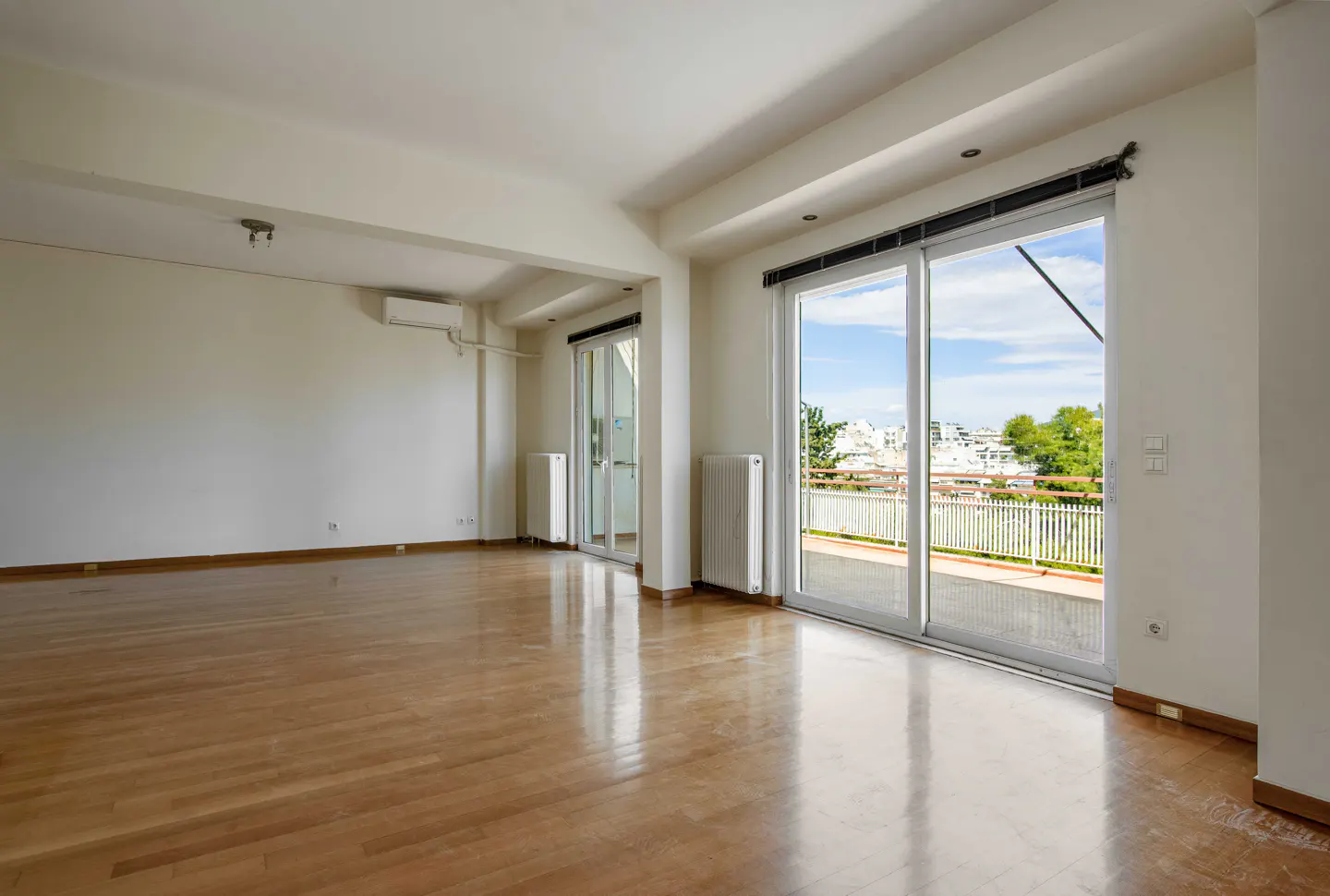 Bright, empty room with hardwood floors, white walls, and sliding glass doors to a balcony with a city view.