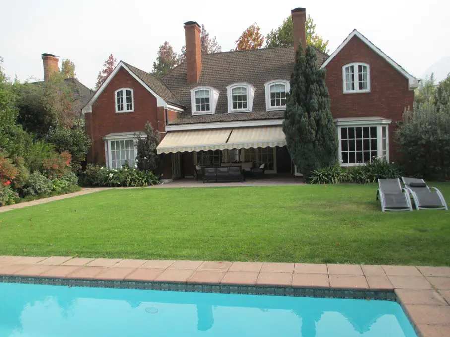 Backyard view of a two-story red brick house with a pool, green lawn, and patio furniture.