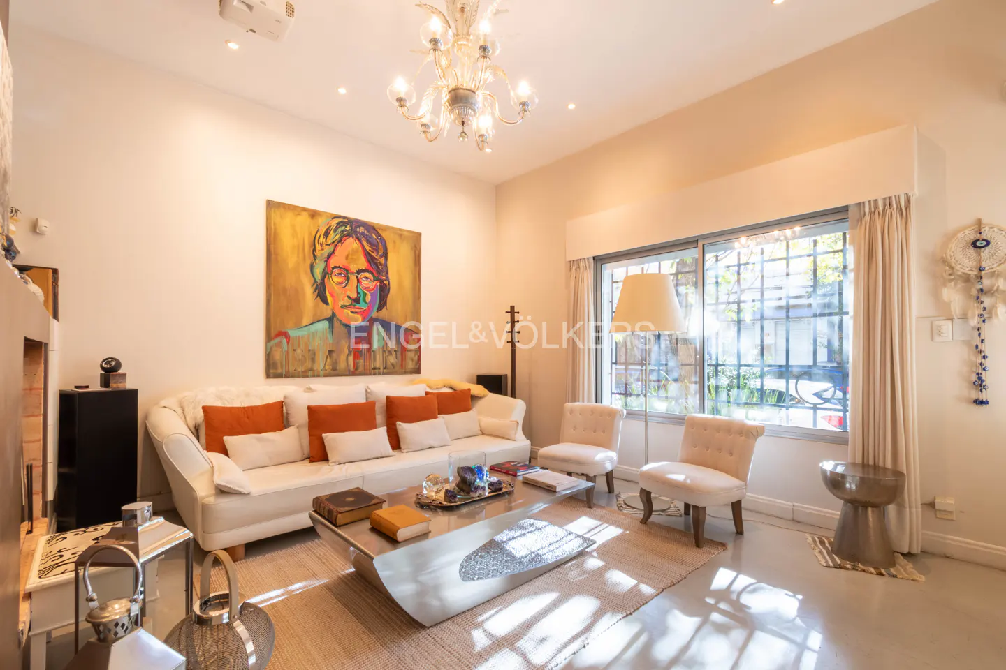 A bright living room with a white sofa, orange pillows, and a large painting of John Lennon. A chandelier hangs from the ceiling.