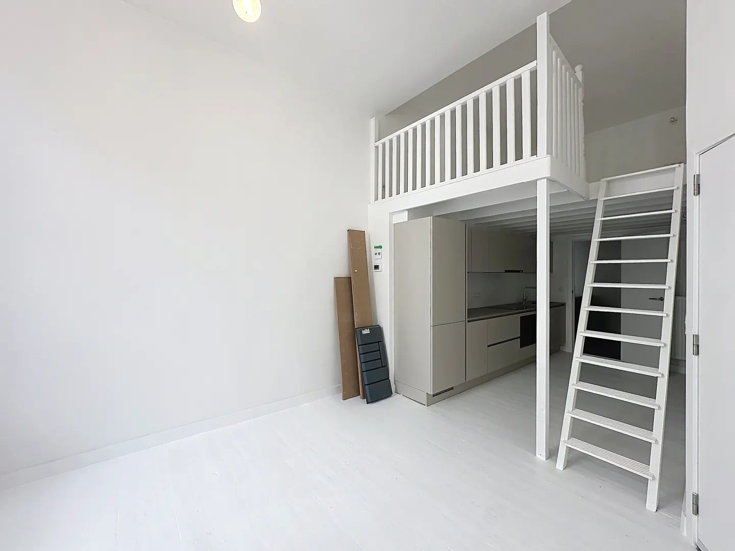 Bright, all-white studio apartment with a loft bed accessed by a ladder. A small kitchen is visible beneath the loft.