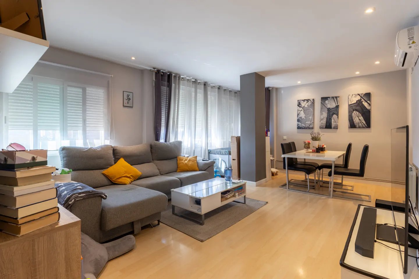 Open-concept living and dining area with a gray sectional sofa, white coffee table, and a dining table with black chairs. Three bridge art pieces hang on the wall.
