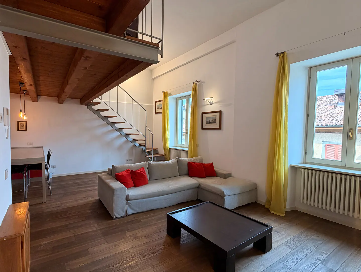 Bright living room with hardwood floors, a gray sectional sofa with red pillows, and a black coffee table. Stairs lead to a loft area with a wooden ceiling. Yellow curtains frame the windows.