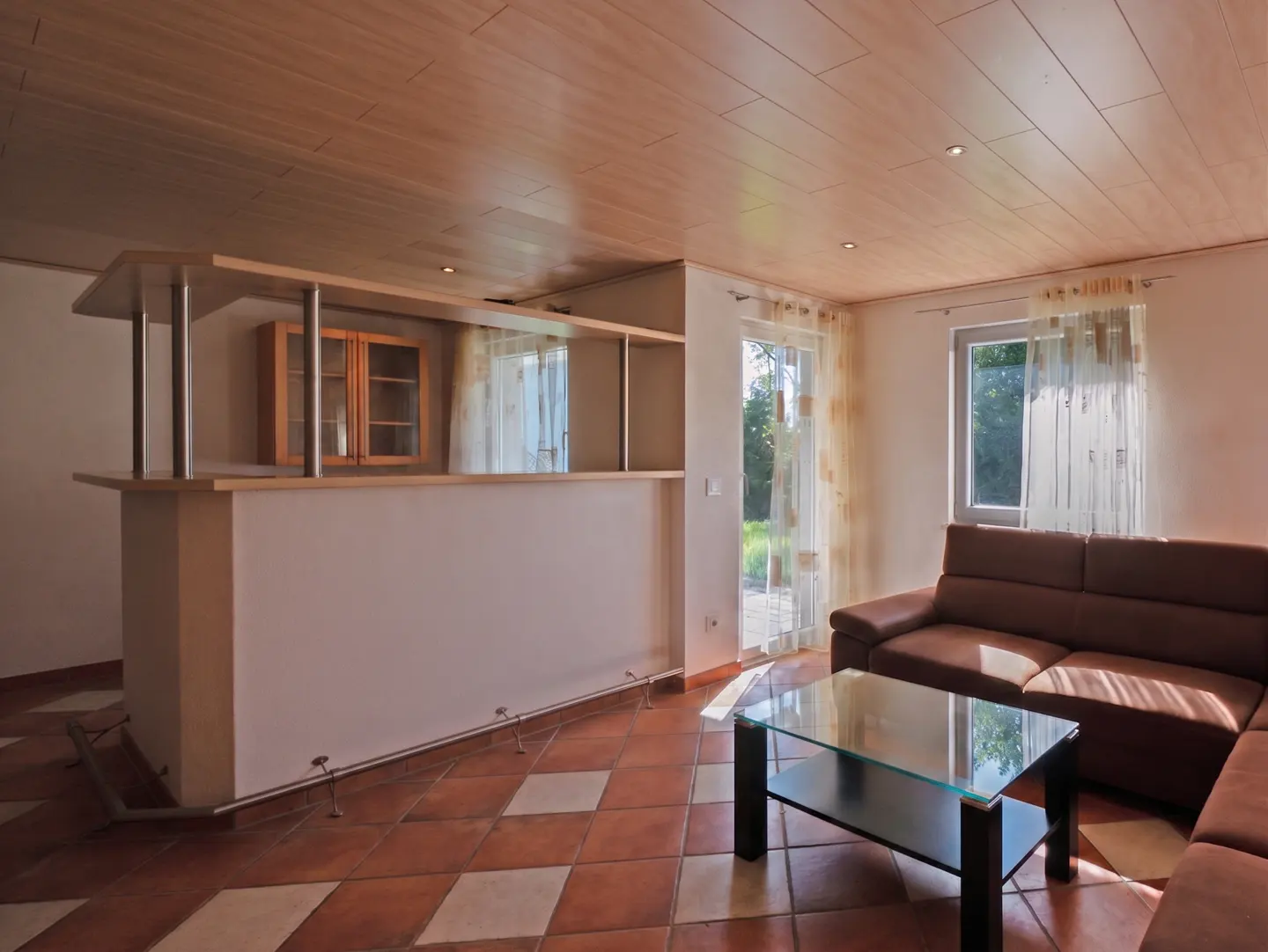 Living room with brown leather sofa, glass coffee table, and bar area with wood cabinets. Tiled floor and wood paneled ceiling.