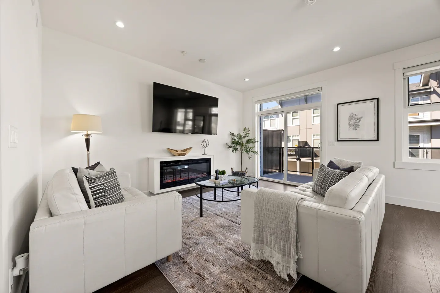 Bright living room with white walls, dark wood floors, and a gray patterned rug. Two white sofas face a TV above an electric fireplace. A glass door leads to a balcony.