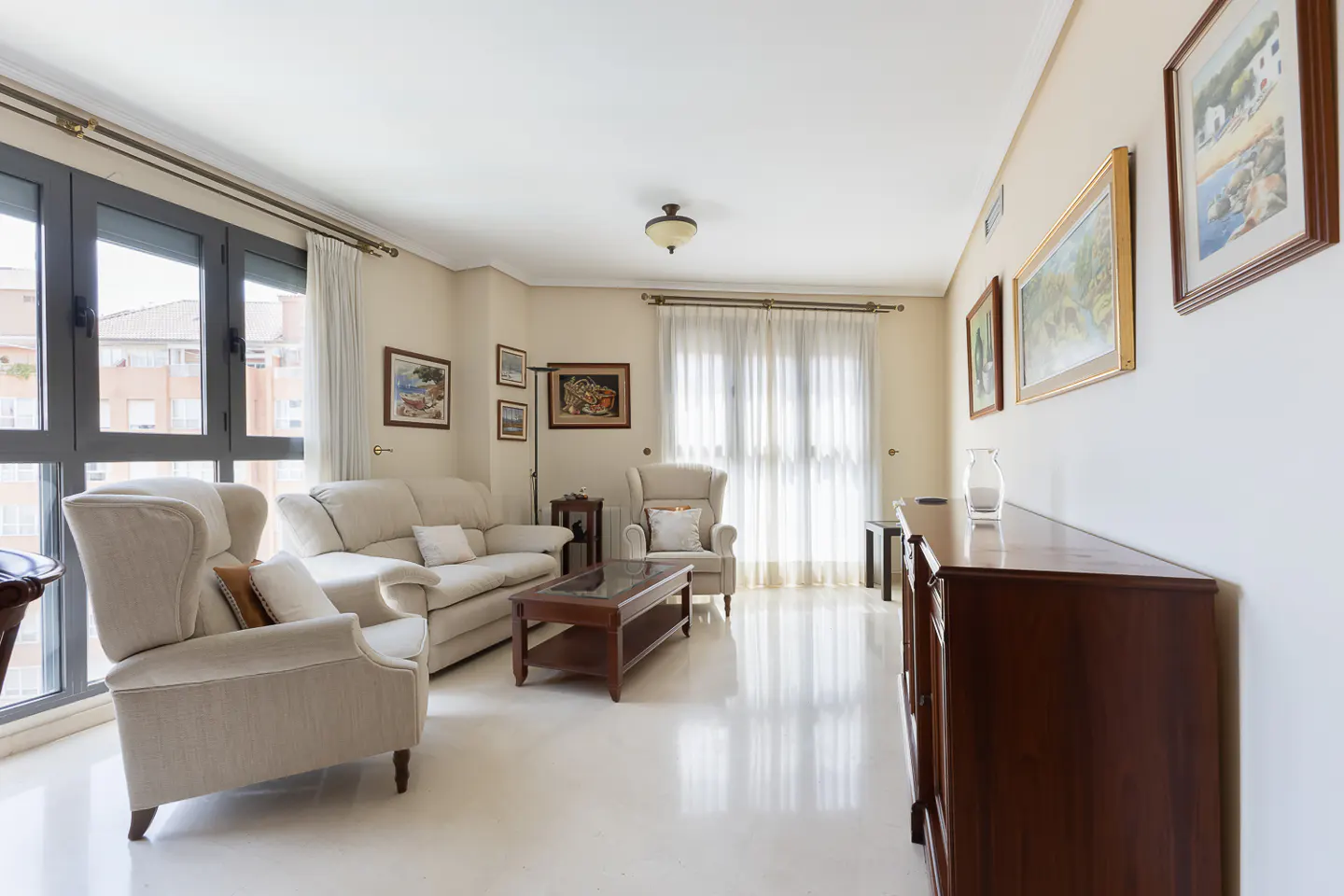Bright living room with beige walls, white tile floor, sofa, armchairs, dark wood furniture, and framed art. Large windows let in natural light.