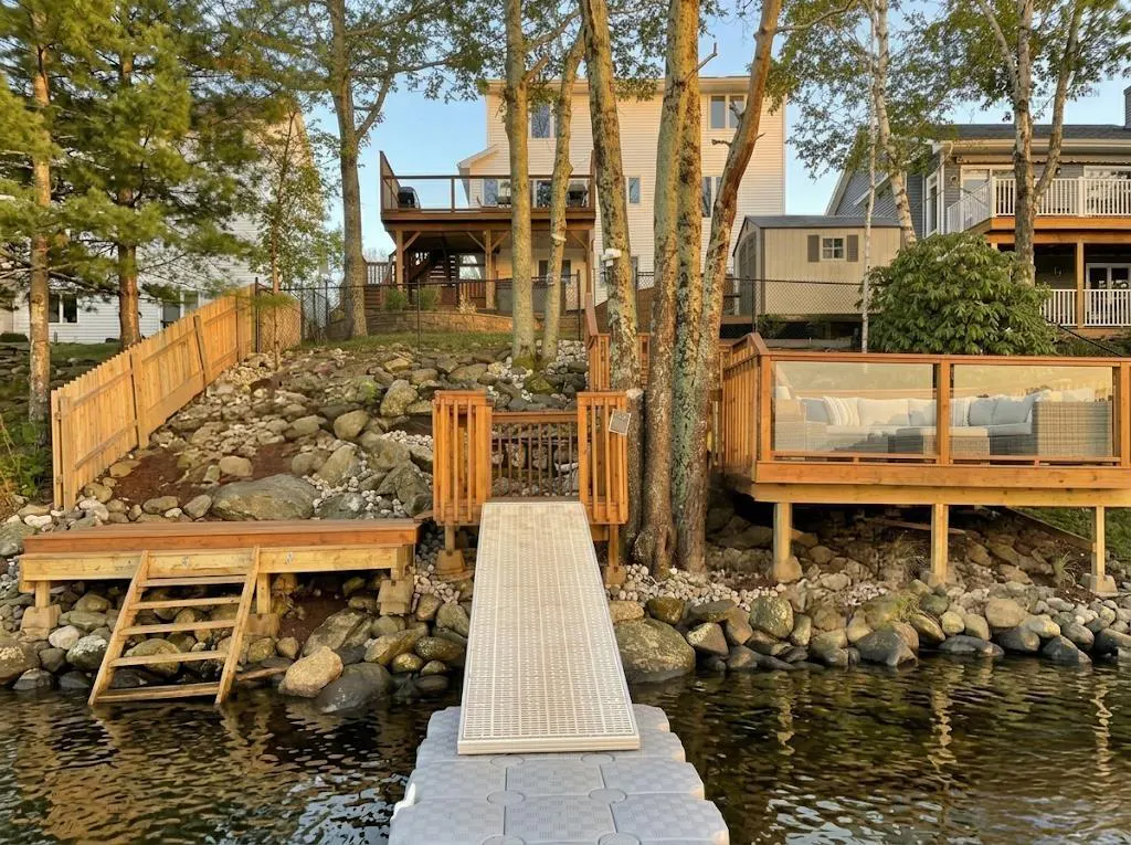 Waterfront property with a dock, decks, and a rocky shoreline. Houses are visible in the background.