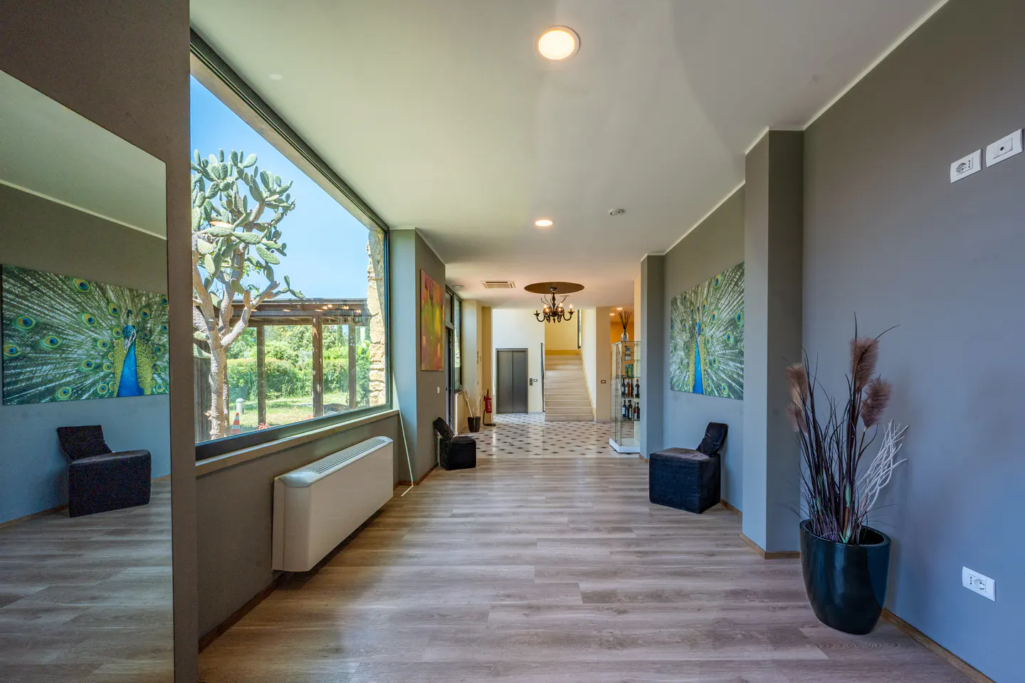 Hallway with gray walls, light wood floors, and large windows. Peacock art, chairs, and a potted plant decorate the space. A staircase and elevator are visible in the background.