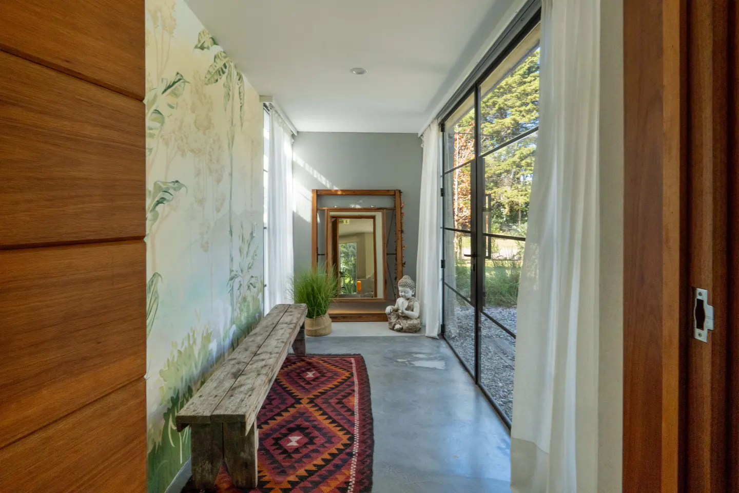 Hallway with a wooden bench, patterned rug, and a Buddha statue near a large window with white curtains. A mirror reflects the outdoors.