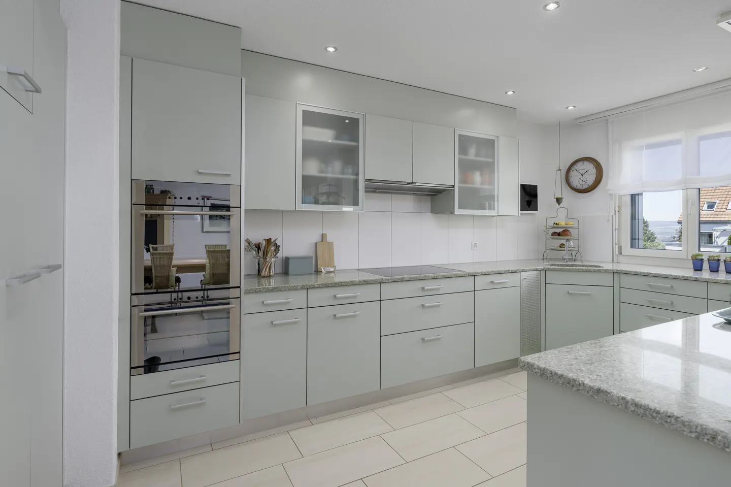Bright kitchen with light green cabinets, granite countertops, and stainless steel appliances. A window with sheer curtains lets in natural light.