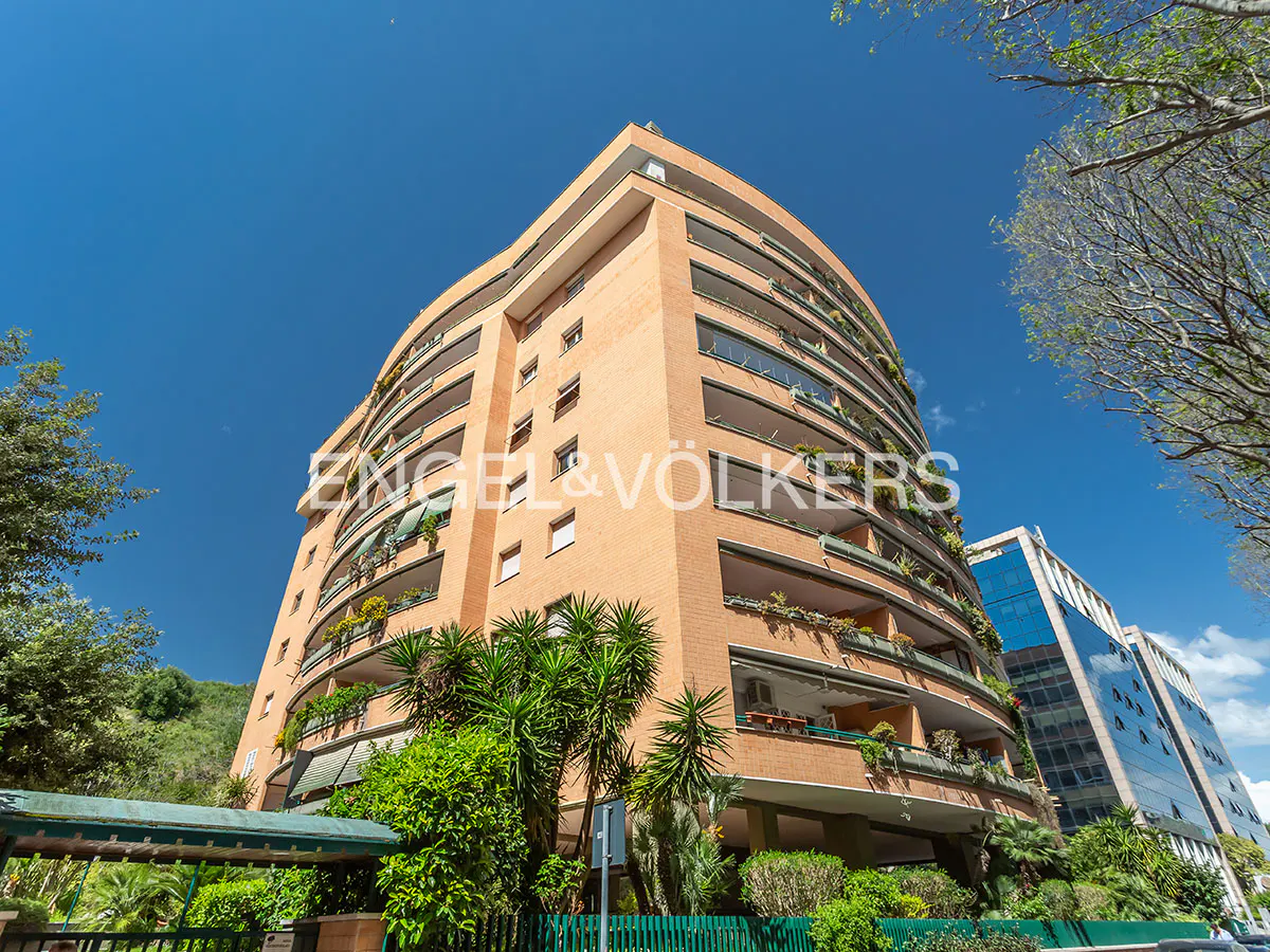 Exterior view of a curved, multi-story brick apartment building with balconies and lush greenery against a clear blue sky.