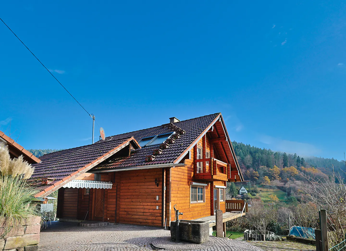 Exterior view of a two-story log cabin with a brown roof, a balcony, and a stone water fountain in the front yard. The background shows a hillside with trees and a clear blue sky.
