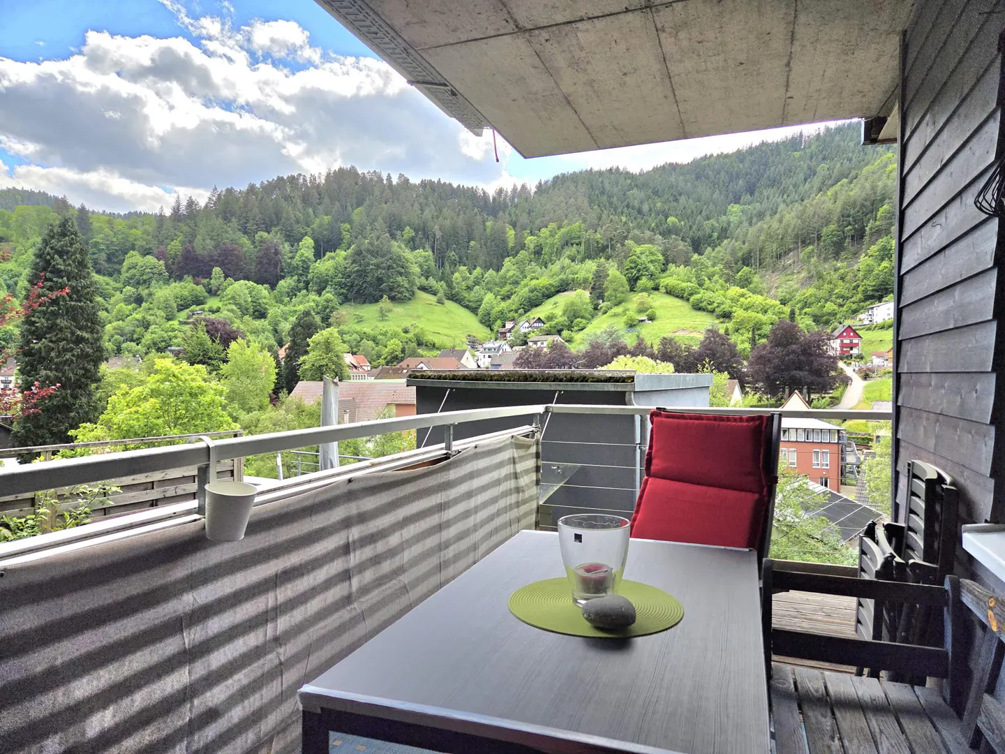 Balcony view of lush green hills. Table with candle, stone, and chair with red cushion. Gray privacy screen and wood siding.