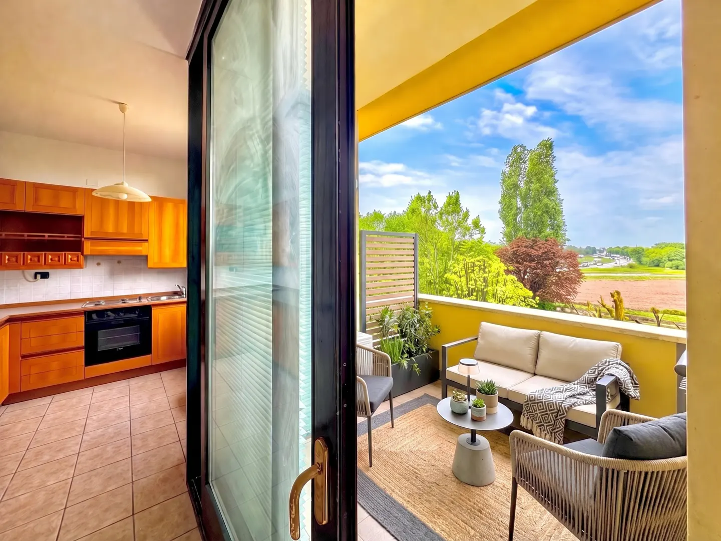 View from kitchen to balcony with outdoor seating, sofa, and green landscape.