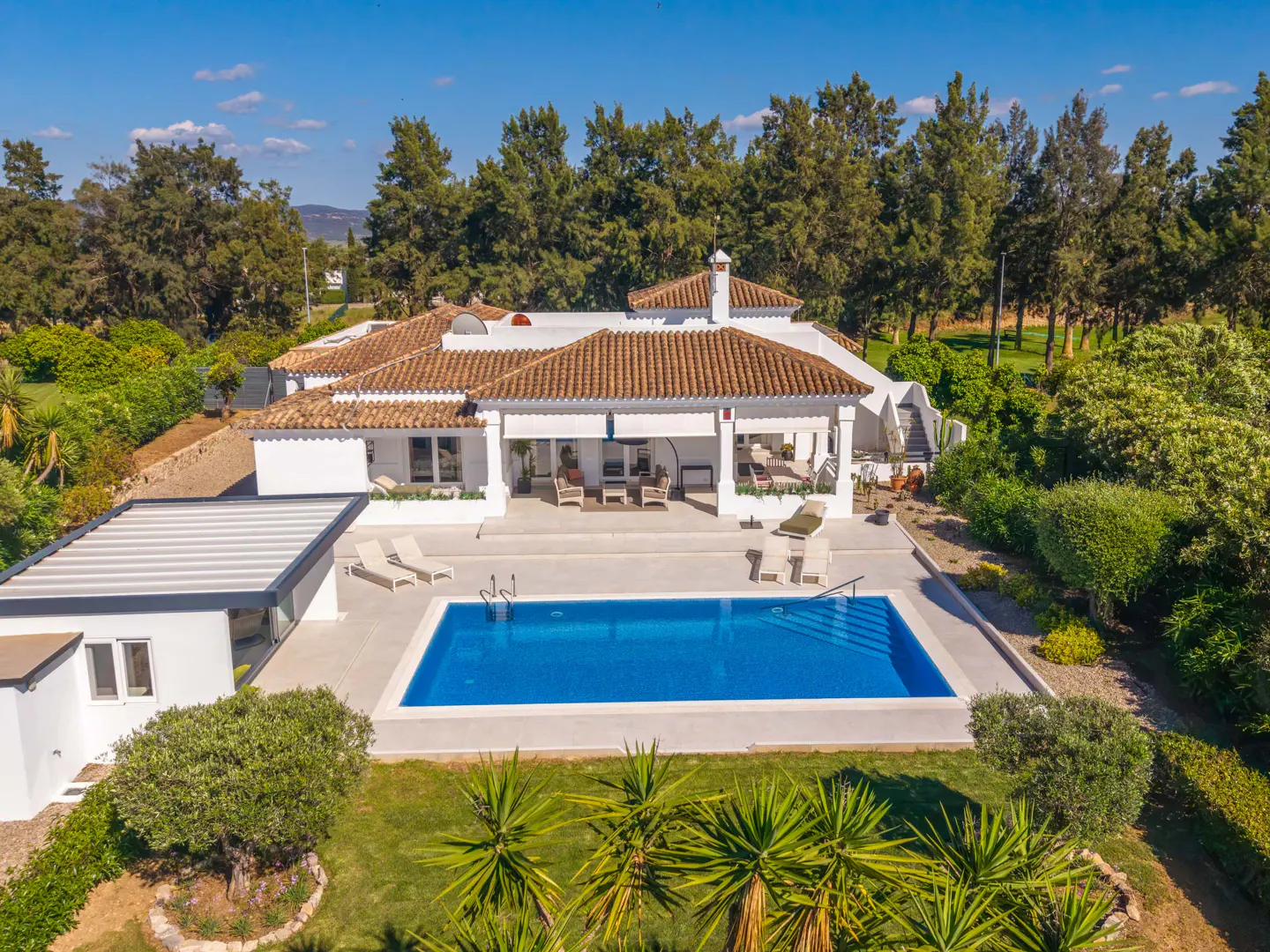 Aerial view of a white villa with a red tile roof, a blue pool, and green trees under a blue sky.