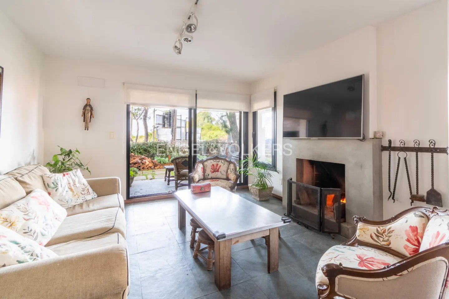 Living room with a beige sofa, floral armchairs, a wooden table, and a fireplace with a TV above it. Sliding glass doors lead to a patio.