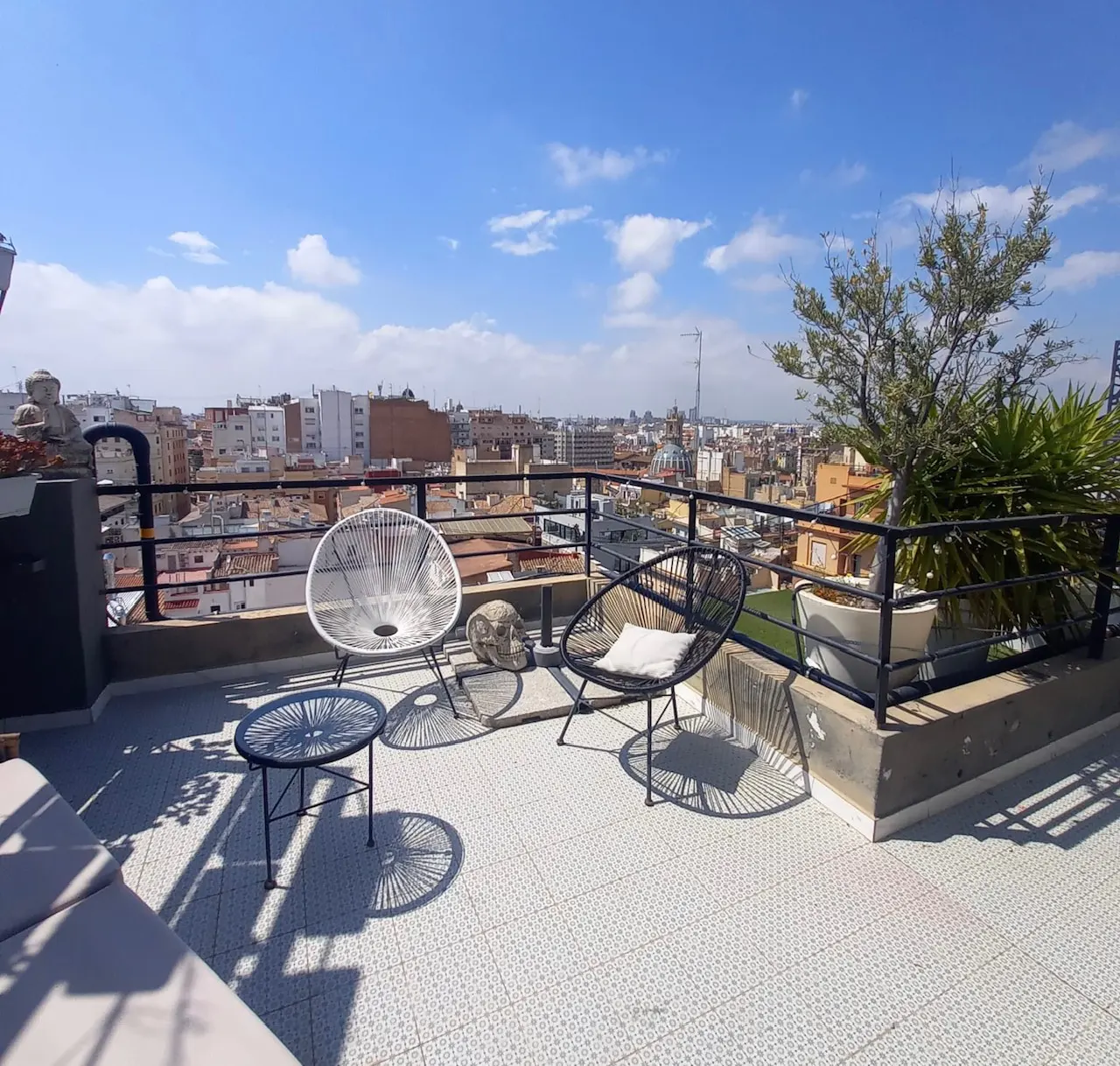 Rooftop patio with white and black chairs, a small table, and potted plants overlooking a city skyline under a blue sky.