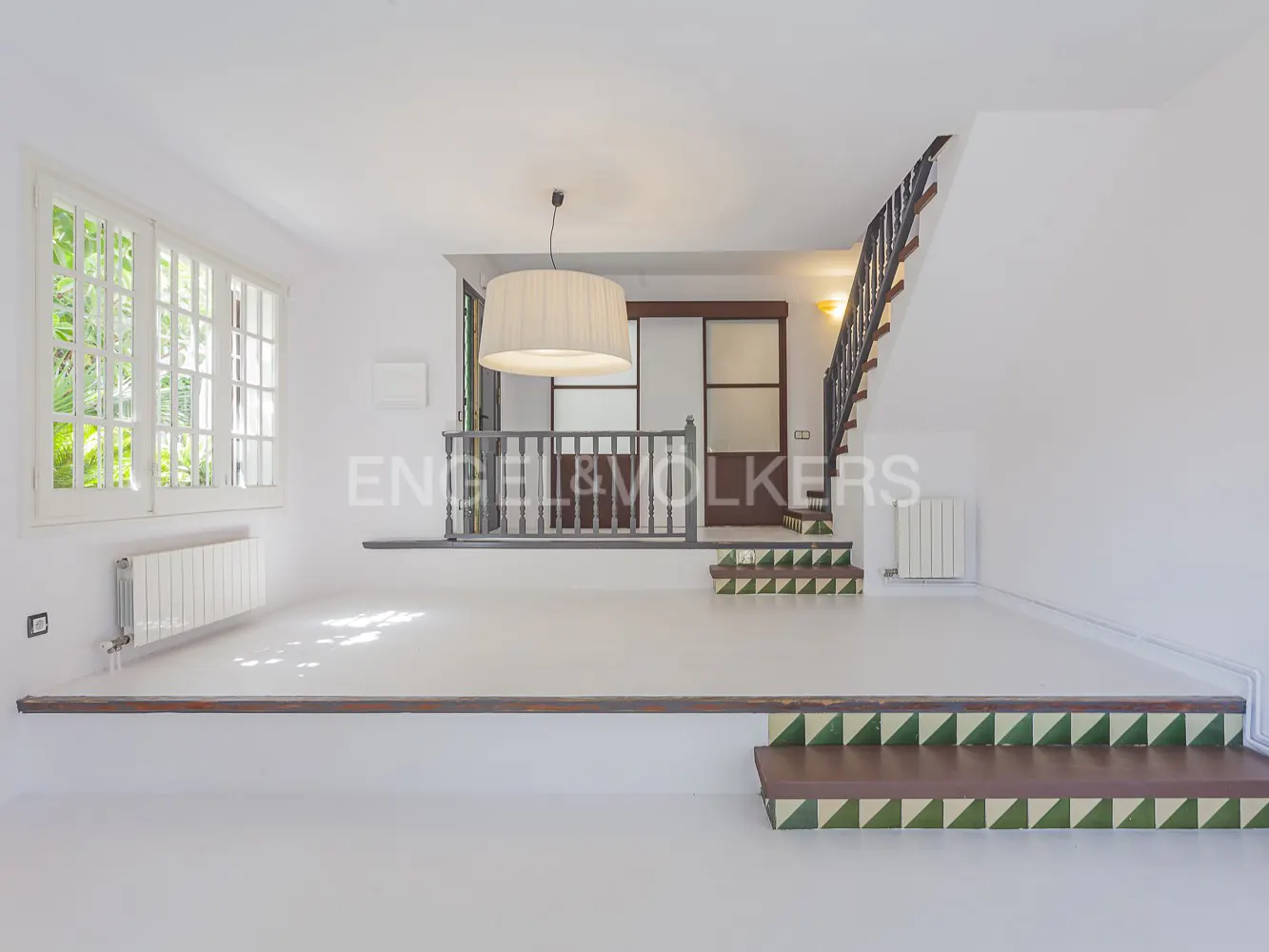Bright, white living room with stairs, a balcony, and a large window. A drum pendant light hangs from the ceiling.