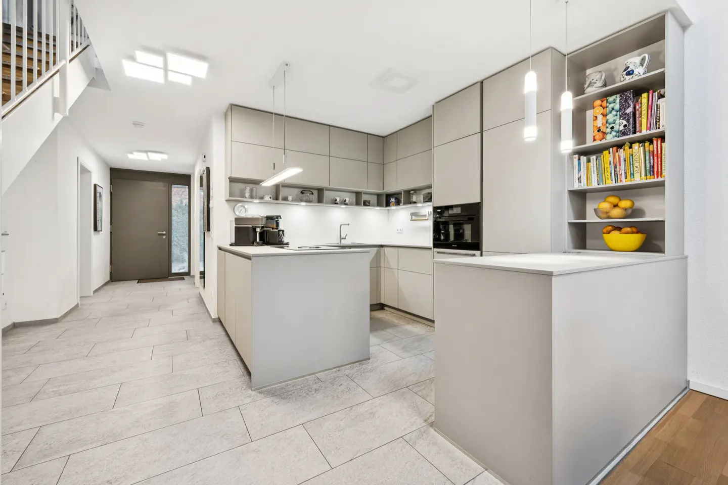 Modern kitchen with gray cabinets, island, and open shelving with books and fruit. Tiled floor and staircase visible.