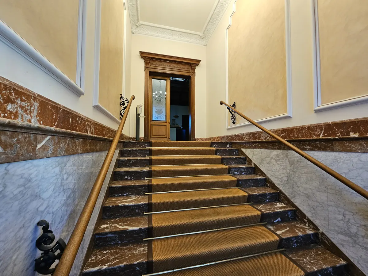 Grand staircase with brown carpet runner, marble steps, and wooden handrails leading to an open wooden door.