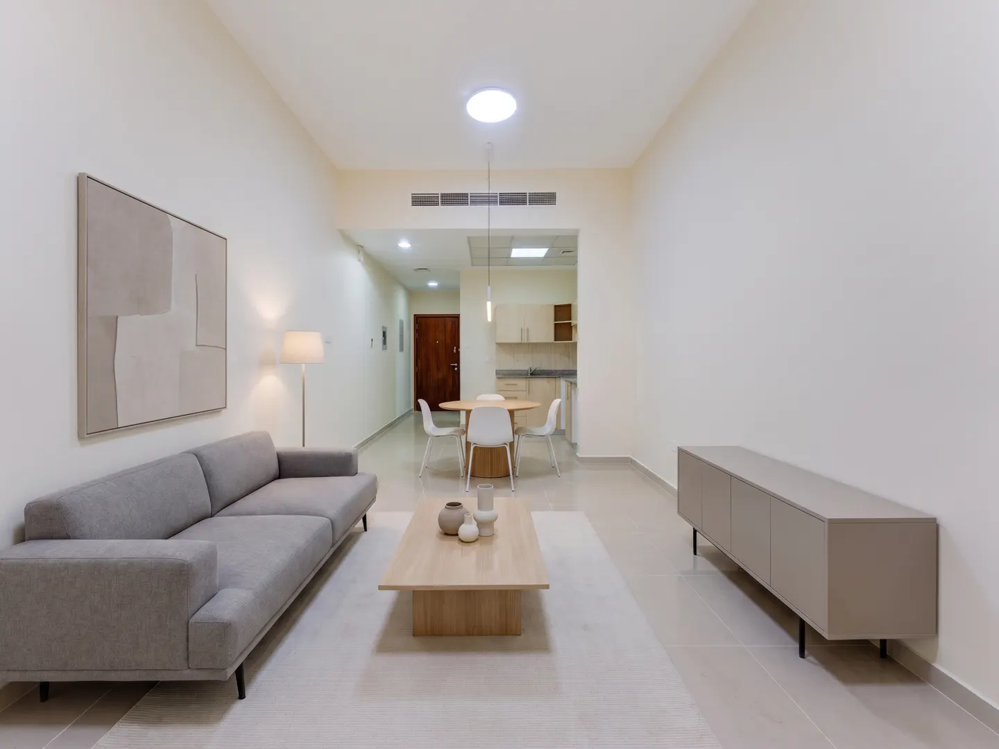 Modern living room with a grey sofa, wooden coffee table, beige sideboard, and a dining area in the background.
