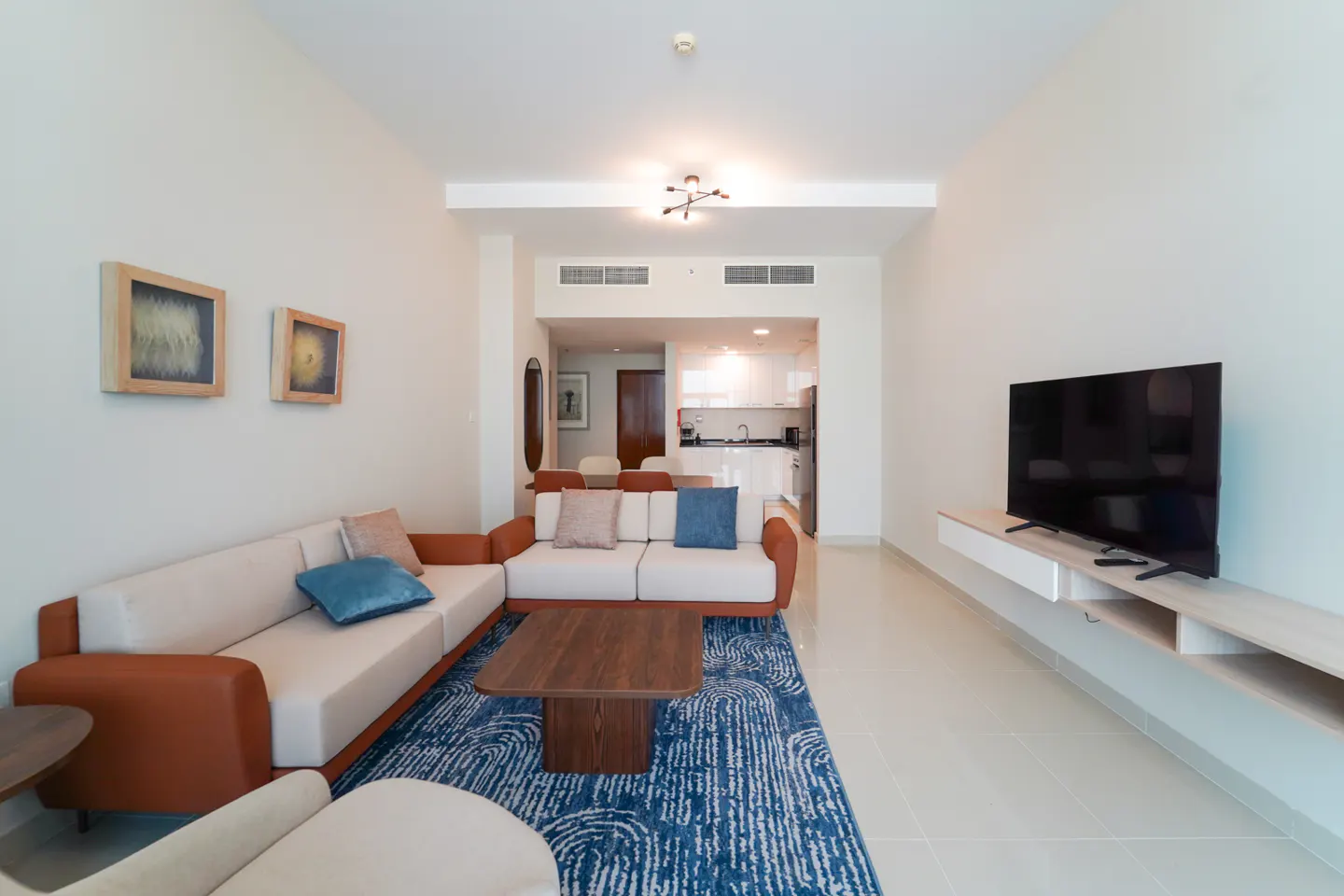 Bright living room with a white and brown sectional sofa, blue rug, and a TV on a white console. A kitchen is visible in the background.