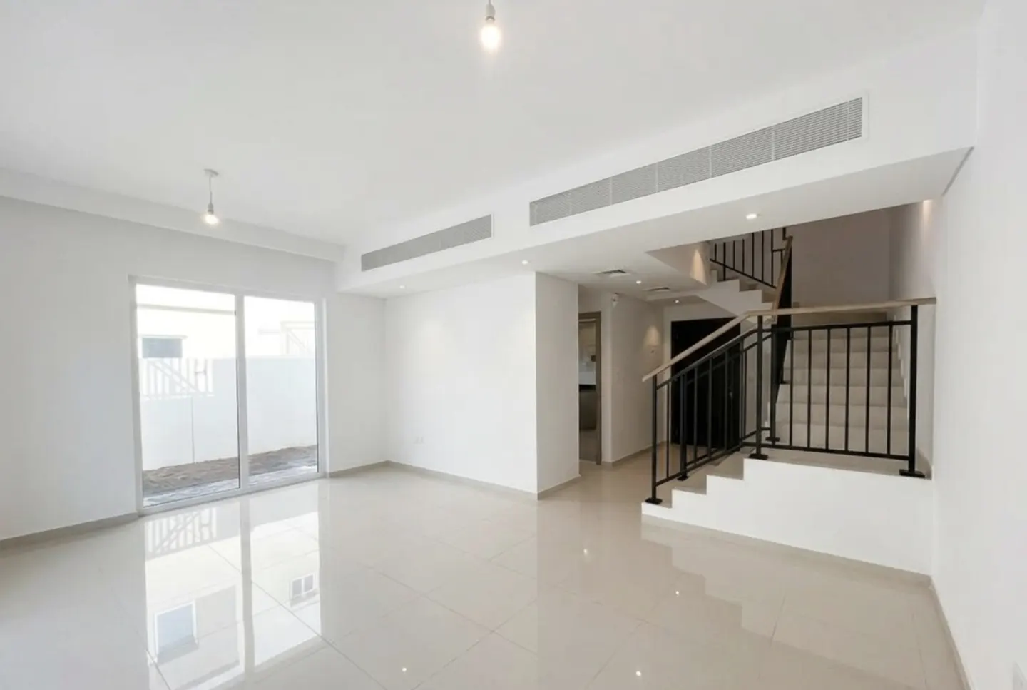 Bright, empty living room with white walls, tile floor, and stairs with black railing. Sliding glass door to the left.