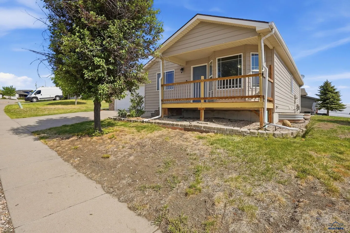 Tan single-story house with a wooden front porch, a small tree in the front yard, and a sidewalk under a blue sky.