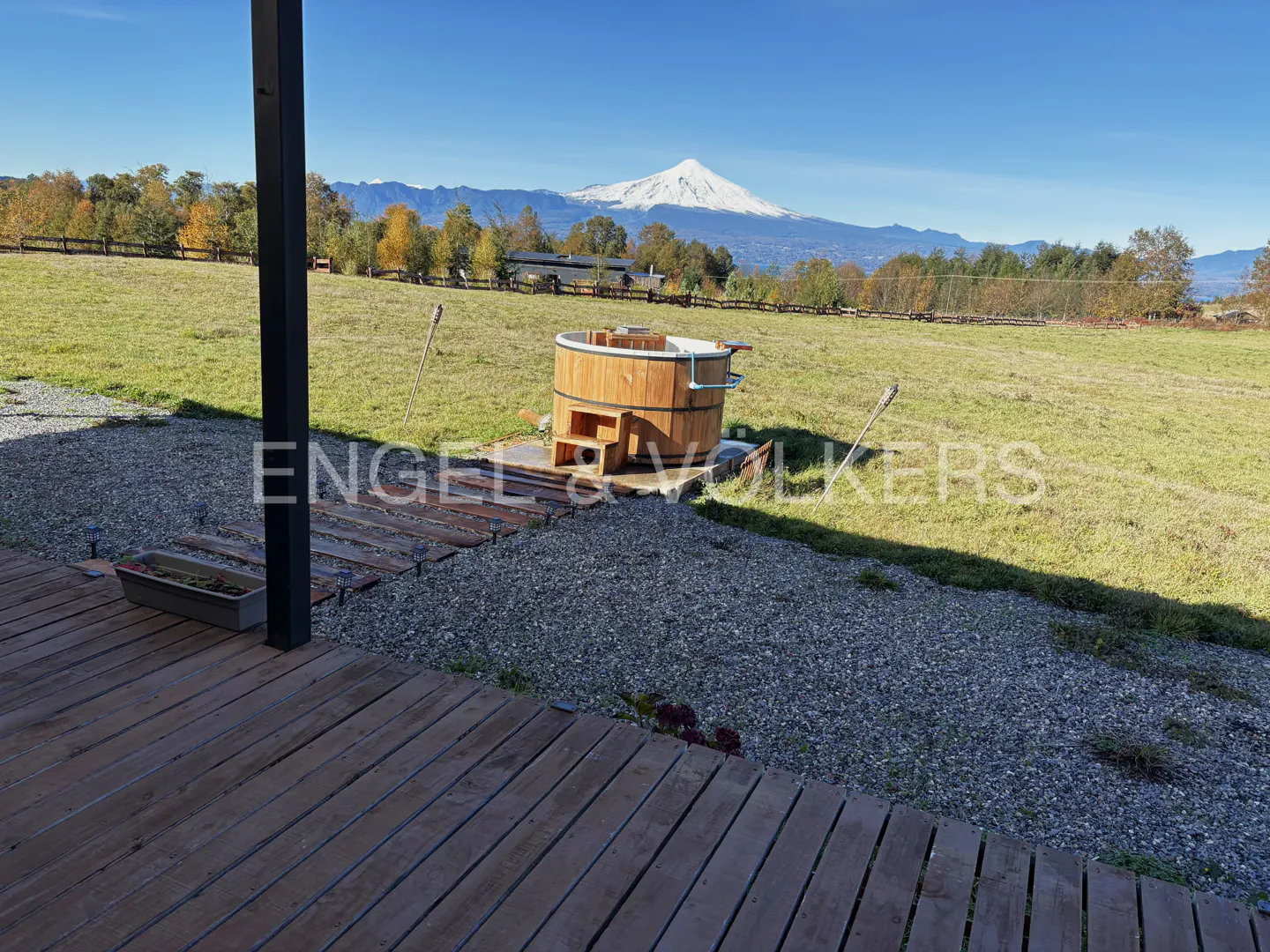 Wooden hot tub on a grassy lawn with a snow-capped mountain in the background, viewed from a wooden deck.