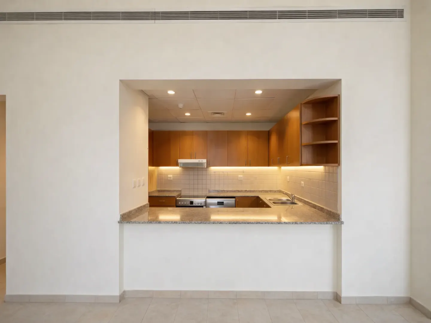 A bright, empty kitchen with wooden cabinets and a marble countertop is viewed through a wall opening.