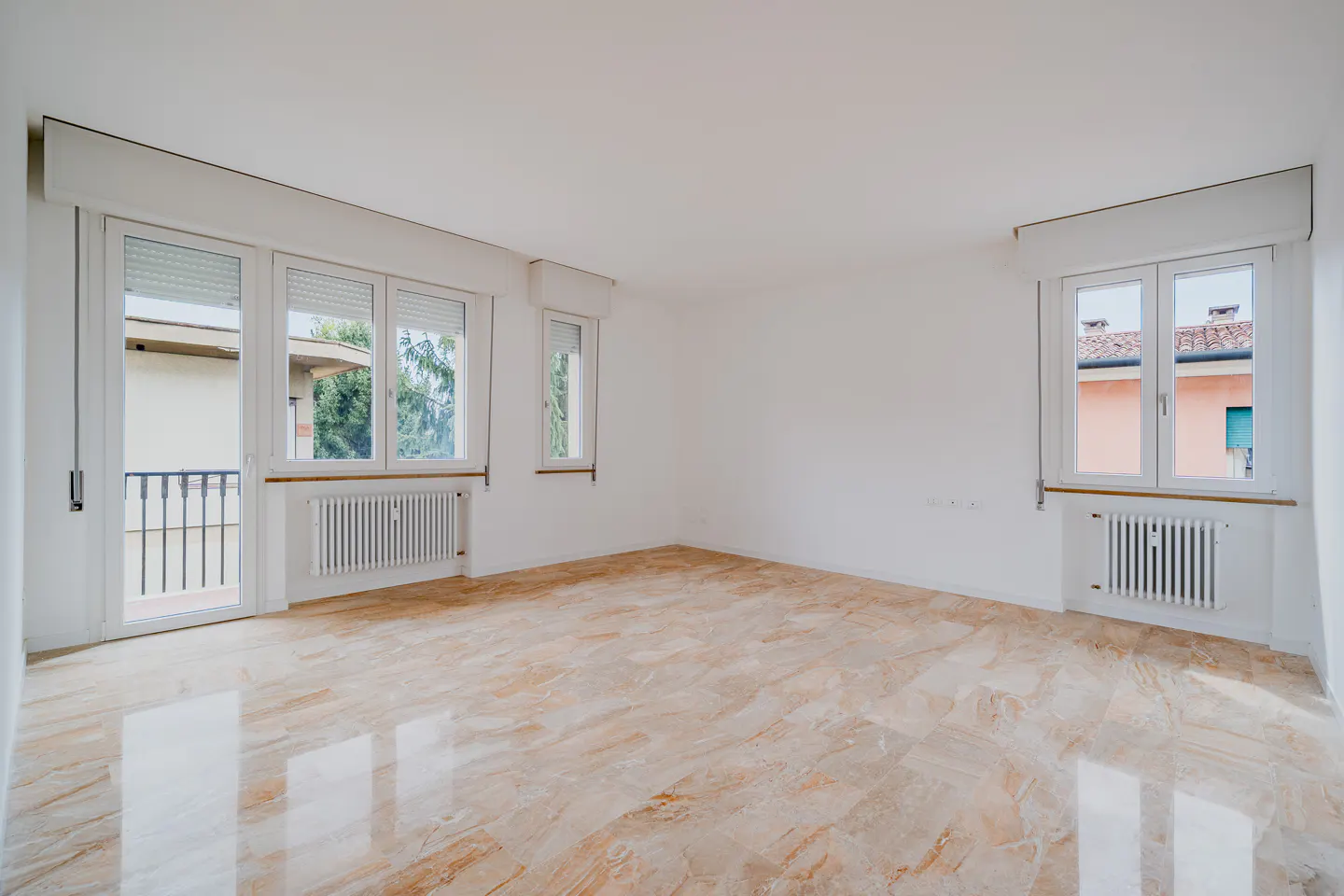 Bright, empty room with white walls, marble floor, and windows. Radiators are under the windows. Balcony door on the left.