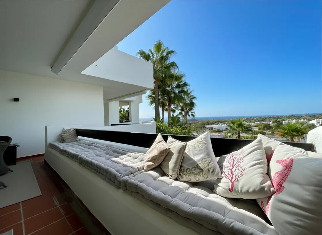 Balcony view with a long white cushioned bench, decorative pillows, and palm trees against a clear blue sky.