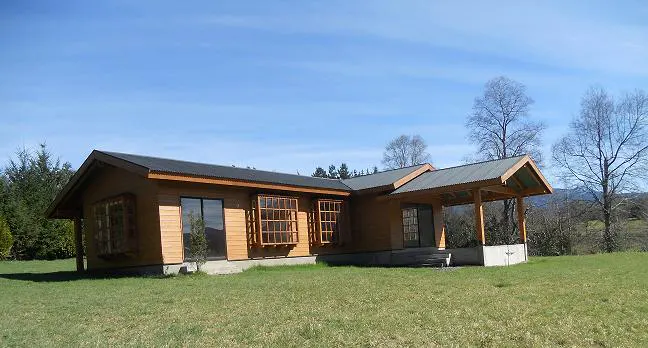 A one-story wood house with a gray roof sits on a green lawn under a blue sky.