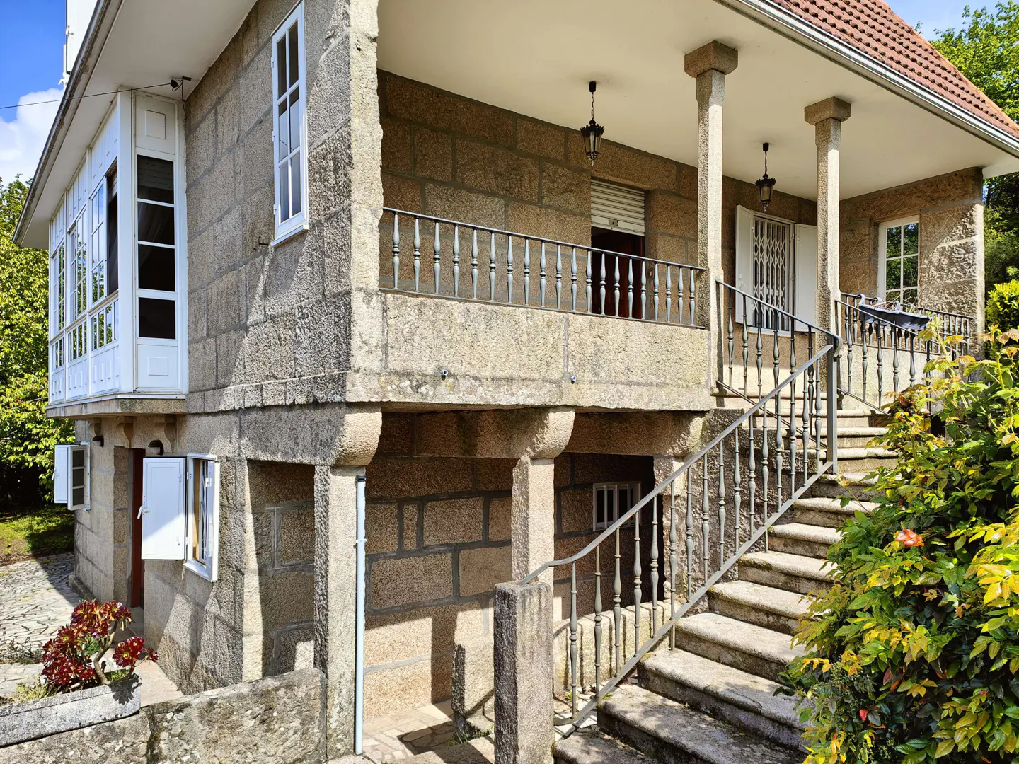 Two-story stone house with a red tile roof, white windows, and a balcony with iron railings. Stone steps lead to the front porch.