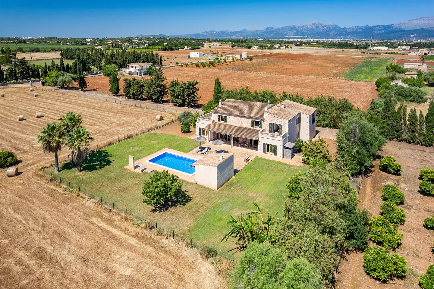 Aerial view of a stone villa with a blue swimming pool, surrounded by expansive rural fields and distant mountains.